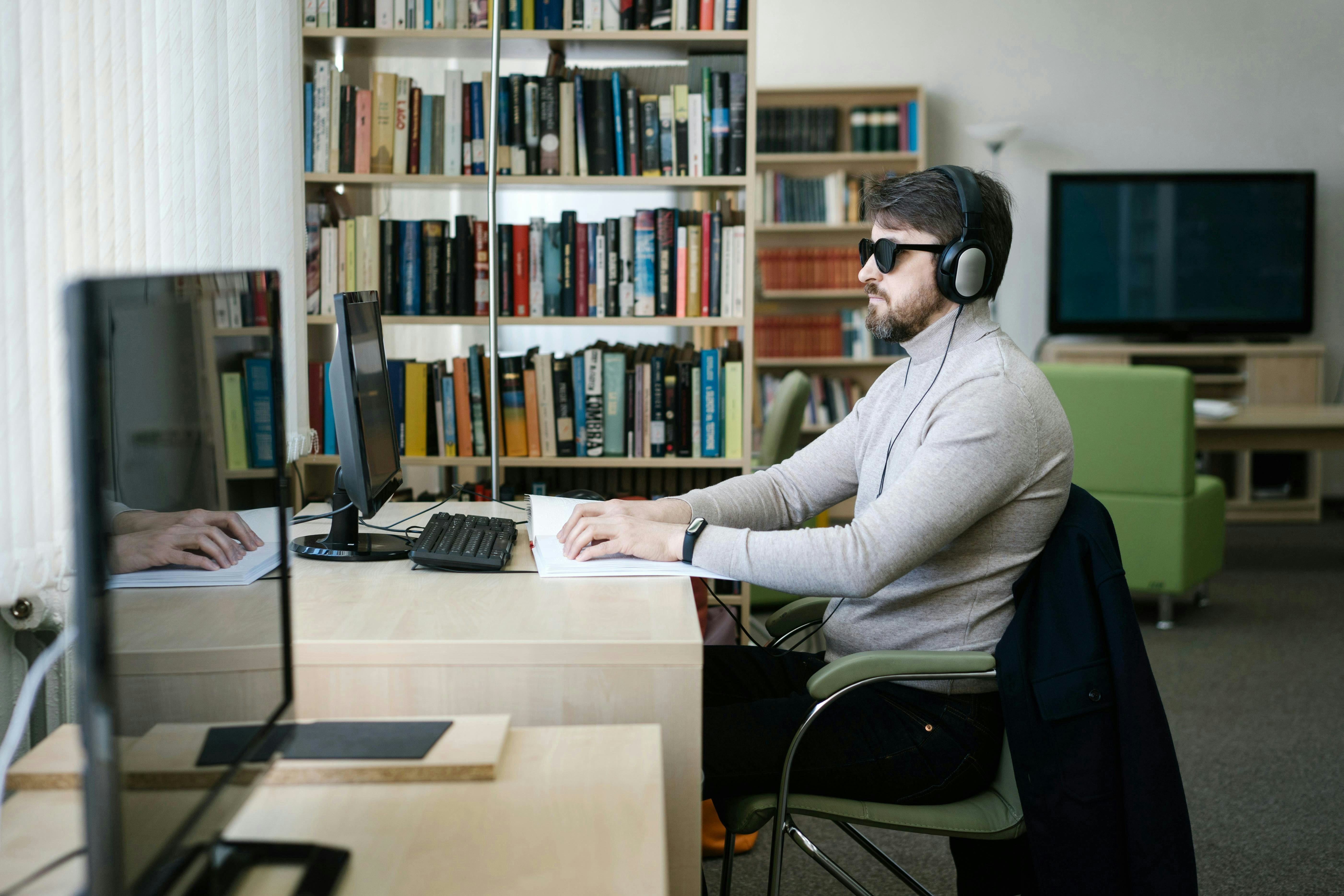 A man wearing headphones and dark glasses uses a braille display at a computer — illustrating how blind users rely on assistive technology to access the web